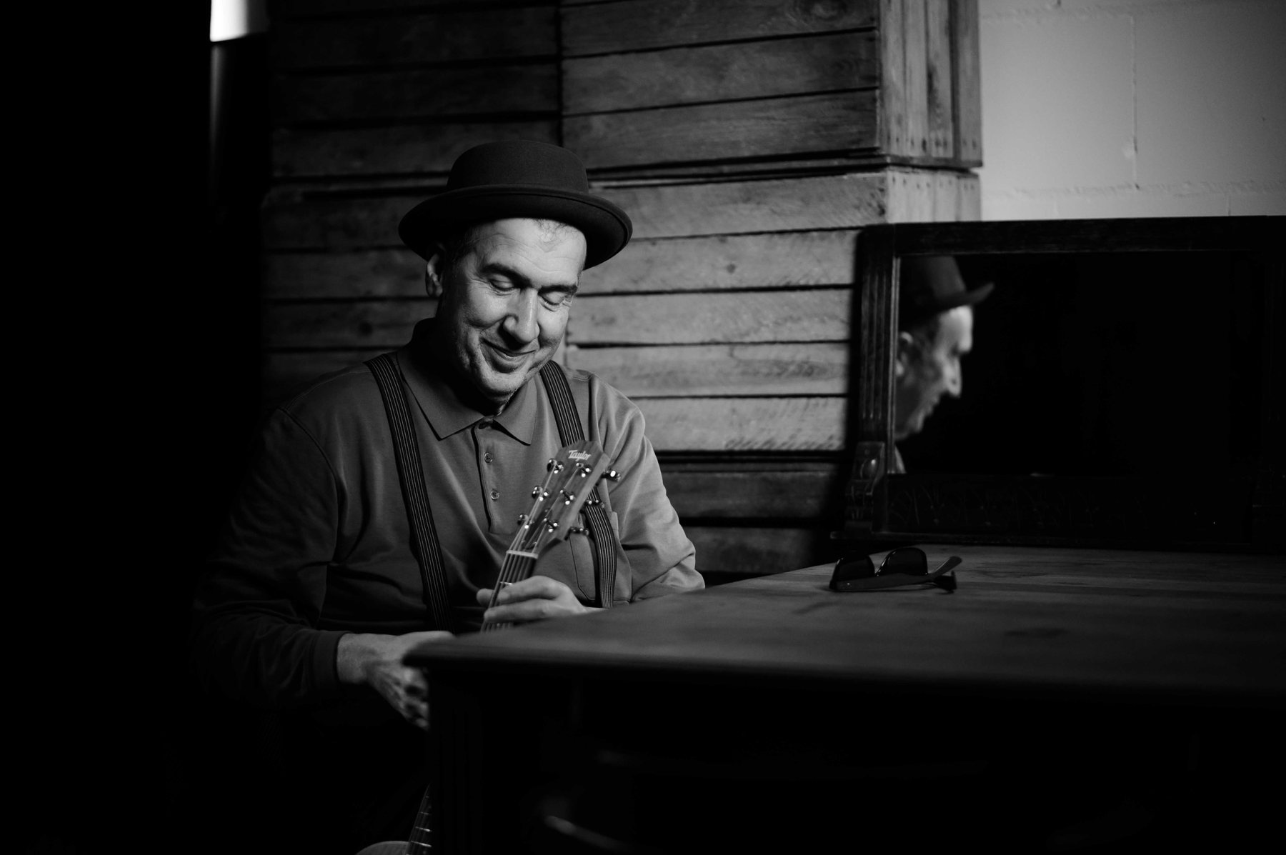 Rick Takvorian sitting at a wooden table with his guitar, smiling, in a dimly lit room.